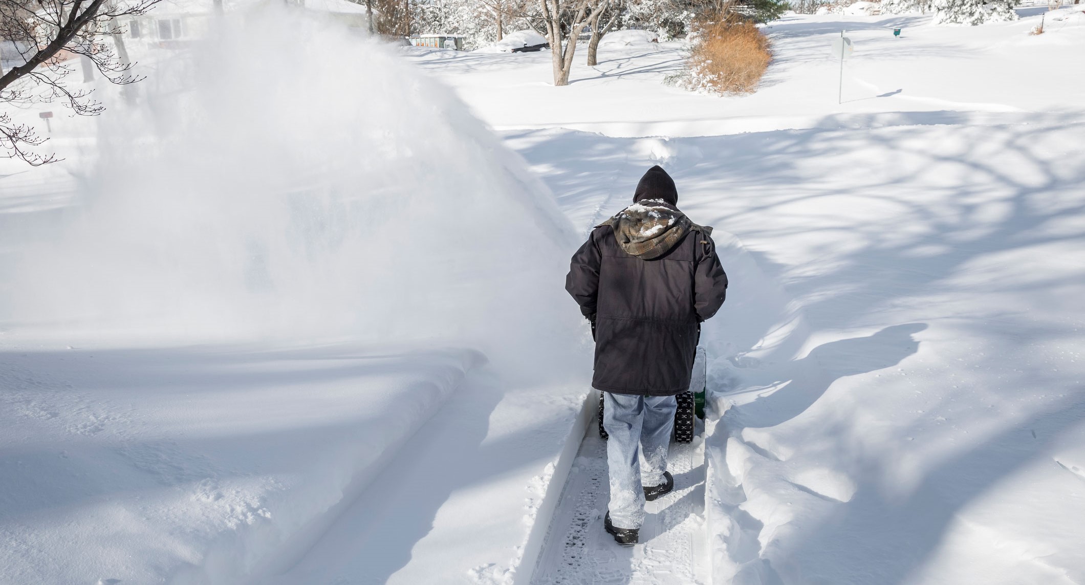 A man from behind pushing a snowblower in a driveway after a blizzard