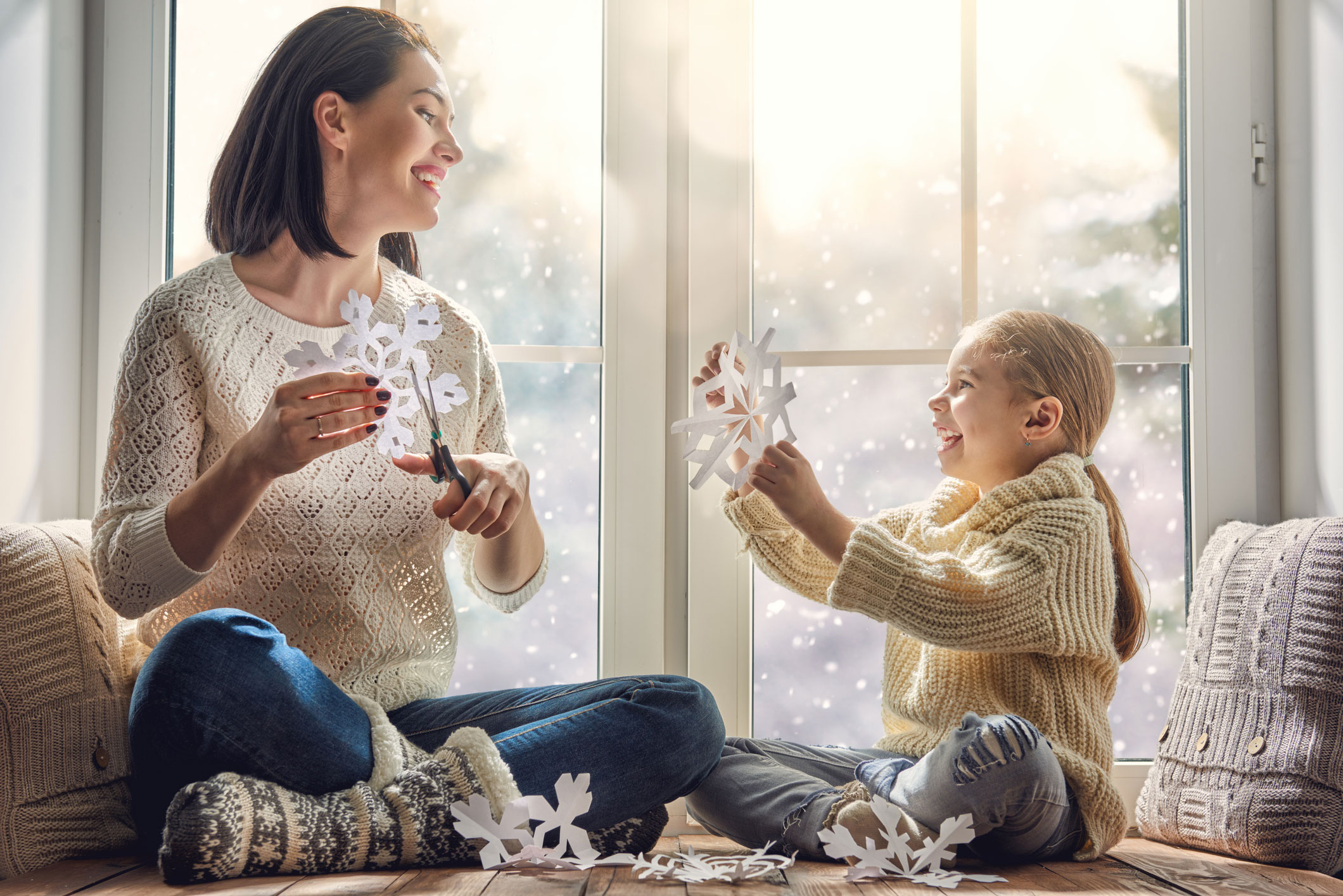 A mom and daughter sitting on a widnow seat making paper snowflakes with a snowy landscape in the background