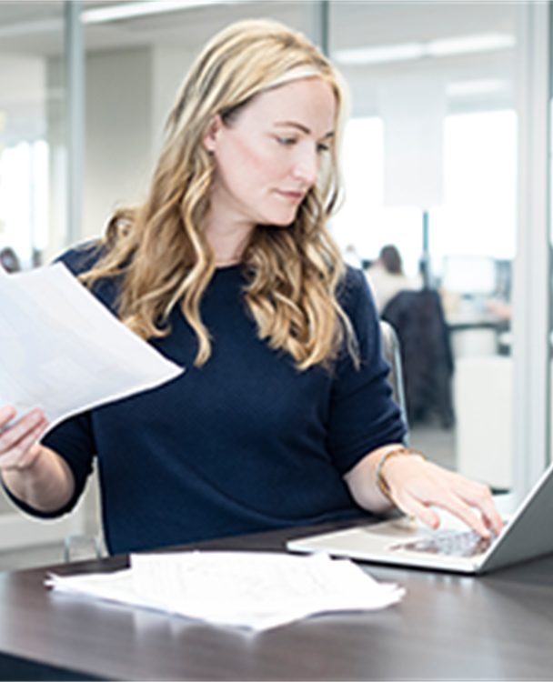 Photo of a blond woman holding a paper and looking at her computer