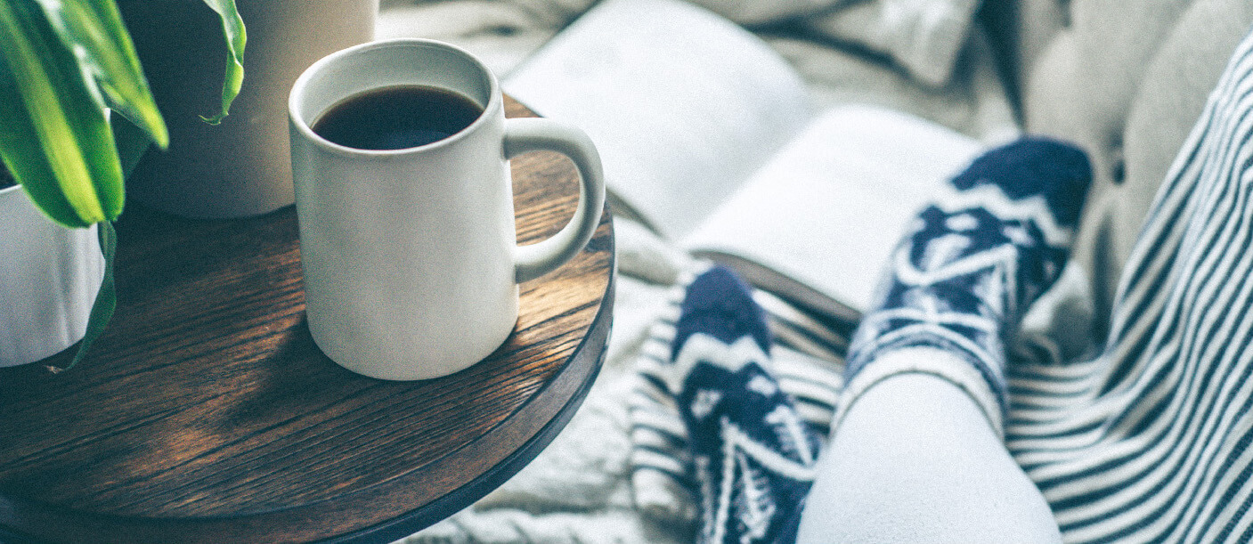 Coffee in mug on wooden table, in front of couch end with socked feet and open book