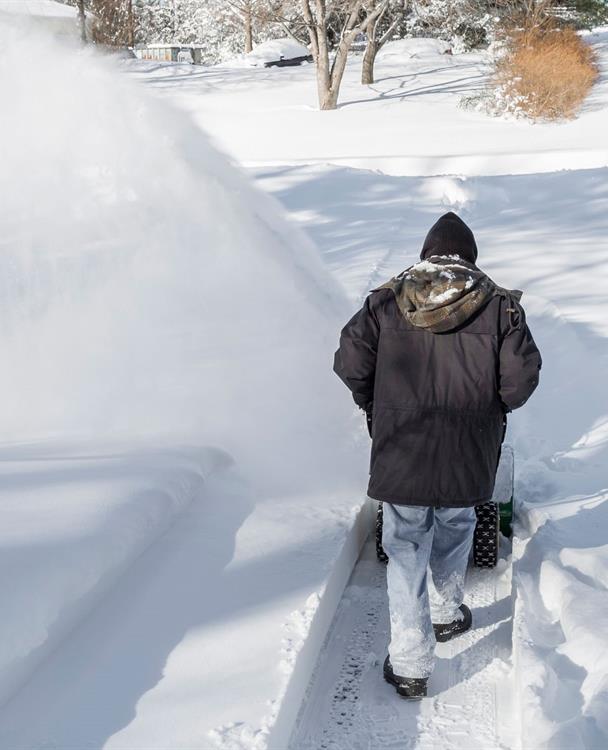 A man from behind pushing a snowblower in a driveway after a blizzard